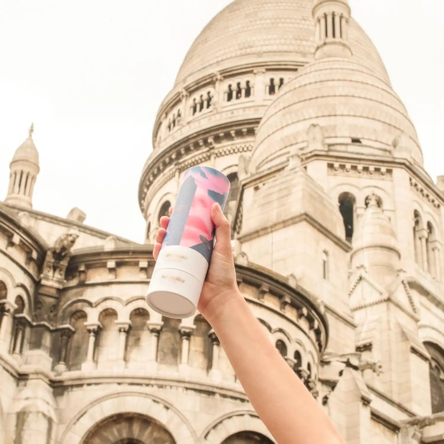 Hand holding a pink and blue beverage can against the Sacré-Cœur Basilica.